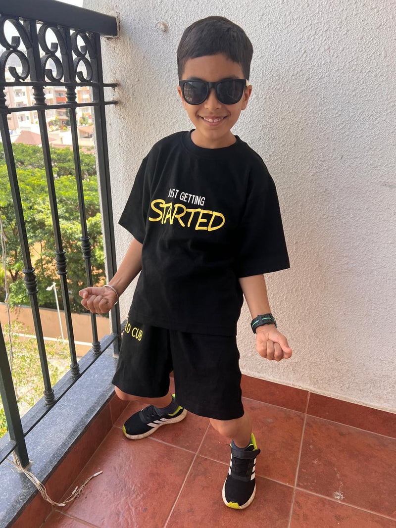A young boy wearing a black t-shirt and matching shorts set with yellow and white text, styled with black sunglasses, standing on a tiled balcony.